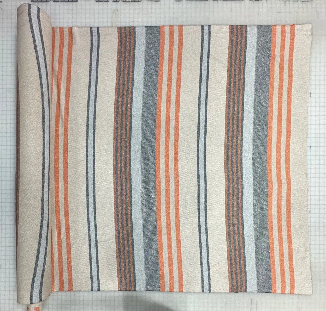 A roll of striped fabric laid flat on a white grid cutting mat. The fabric has vertical stripes in orange, gray, dark blue, and cream.