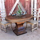 A round wooden dining table with a pedestal base and a distressed metal band around the tabletop, styled with a plant and antlers, flanked by two rustic wooden chairs against a weathered red barn backdrop.