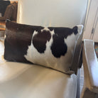 A rectangular cowhide lumbar pillow with a black and white spotted pattern, resting on a cream-colored leather armchair.