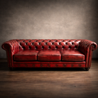 A classic red leather Chesterfield sofa with deep button tufting, rolled arms, and nailhead trim, set against a textured gray wall and dark wood floor.