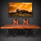 A framed wall art print depicting two bison at sunset hangs above a rustic copper and iron dining table with two brown leather chairs against a dark gray wall.