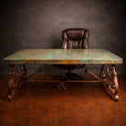 A rustic conference desk with a distressed copper patina top and ornate forged iron legs, paired with a dark brown leather office chair against a dark textured studio backdrop.