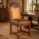 A rustic wooden dining chair with a brown leather seat and back, a tan leather center panel, and dark carved wood arms, shown in a warm, traditional dining room setting next to a wooden table.