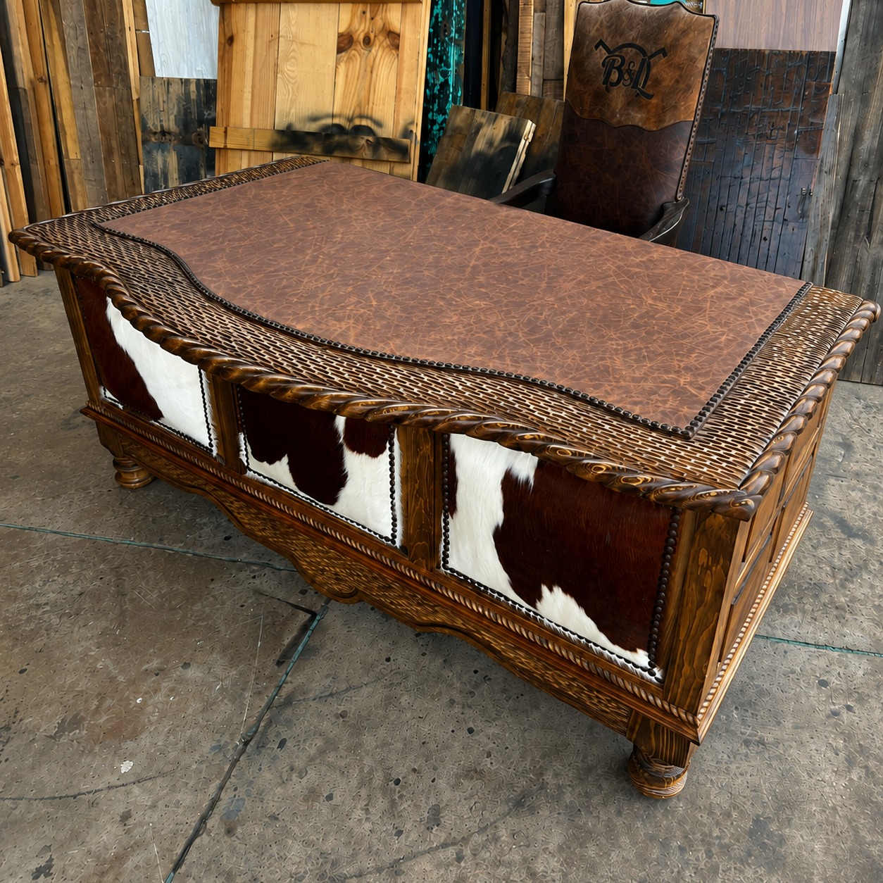 A rustic western-style wooden desk with a brown leather top, carved rope trim, nailhead trim, and three cowhide panel accents on the front, photographed in a workshop or barn setting
