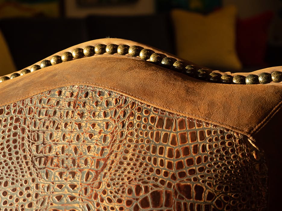 Close-up of a brown textured leather chair back featuring a crocodile pattern and a row of antiqued nailhead trim along the curved top edge.