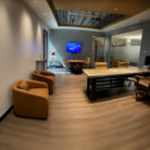 A modern office meeting room with two tan armchairs in the foreground, a large wood-top conference table, office chairs, a mounted TV on a gray wall, glass partitions, and light wood flooring.