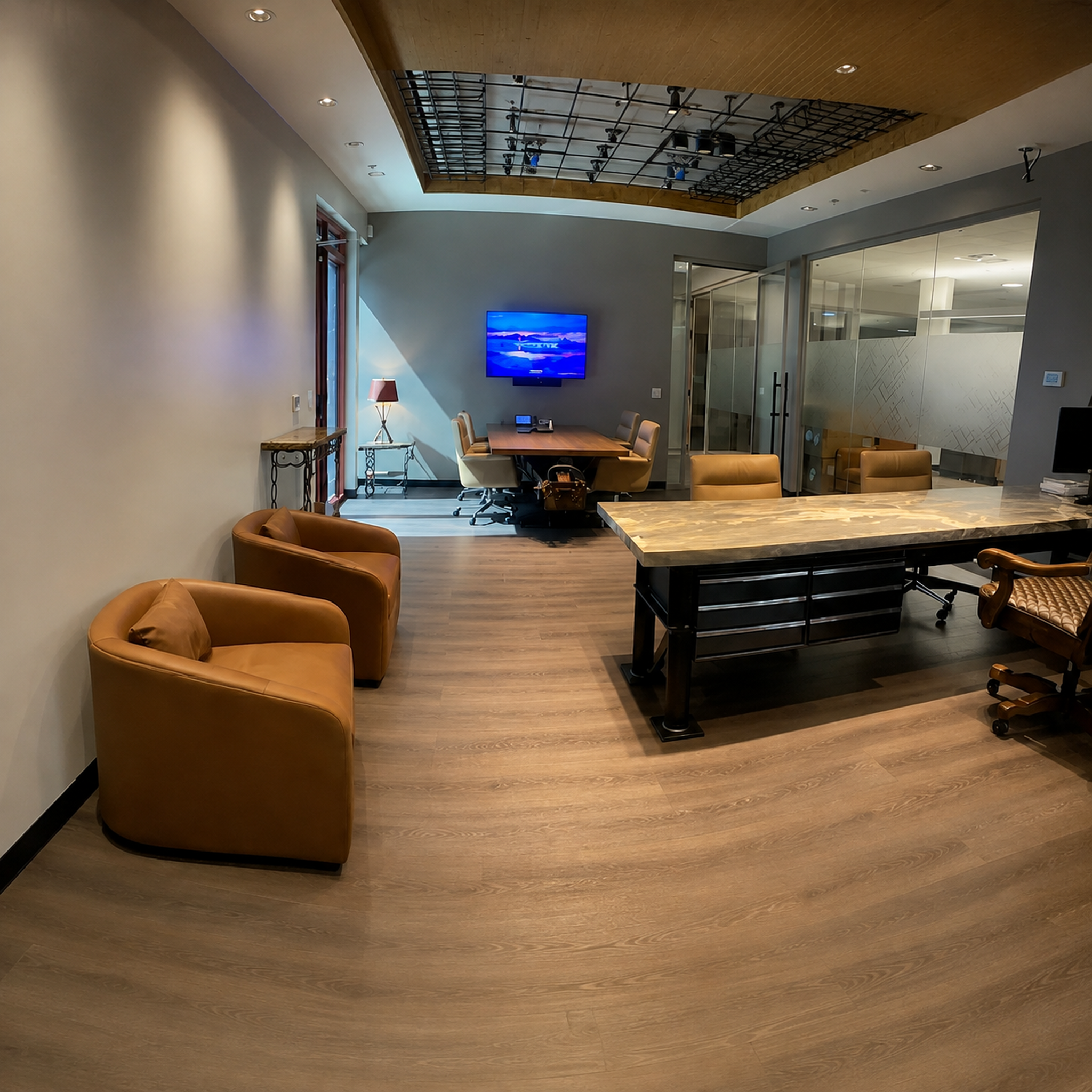 A modern office meeting room with two tan armchairs in the foreground, a large wood-top conference table, office chairs, a mounted TV on a gray wall, glass partitions, and light wood flooring.