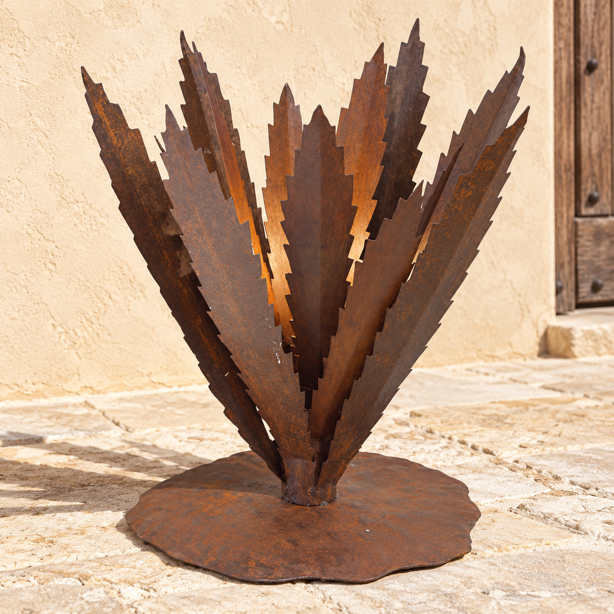 Rust metal garden sculpture shaped like a stylized succulent or palm frond, mounted on a round base, photographed outdoors against a beige stucco wall and stone floor.