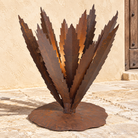 Rust metal garden sculpture shaped like a stylized succulent or palm frond, mounted on a round base, photographed outdoors against a beige stucco wall and stone floor.
