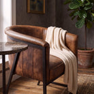 A brown leather armchair with a dark metal frame, styled with a cream knit throw blanket, set in a warm, rustic room next to a marble-top side table and a potted plant.