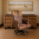 A rustic brown leather and Axis deer-hide swivel office chair positioned in front of a matching dark wood desk, with a framed landscape print of a pronghorn on the beige wall behind.