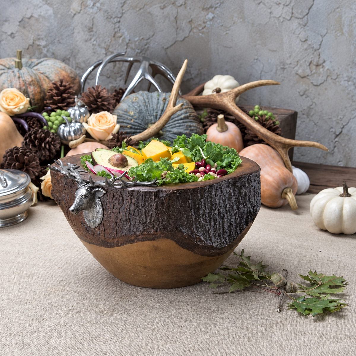 A rustic wooden bowl with a tree trunk design and a pewter bull elk head handle, filled with autumnal vegetables and fruit, set on a beige tablecloth with pumpkins, pinecones, and antlers in the background.