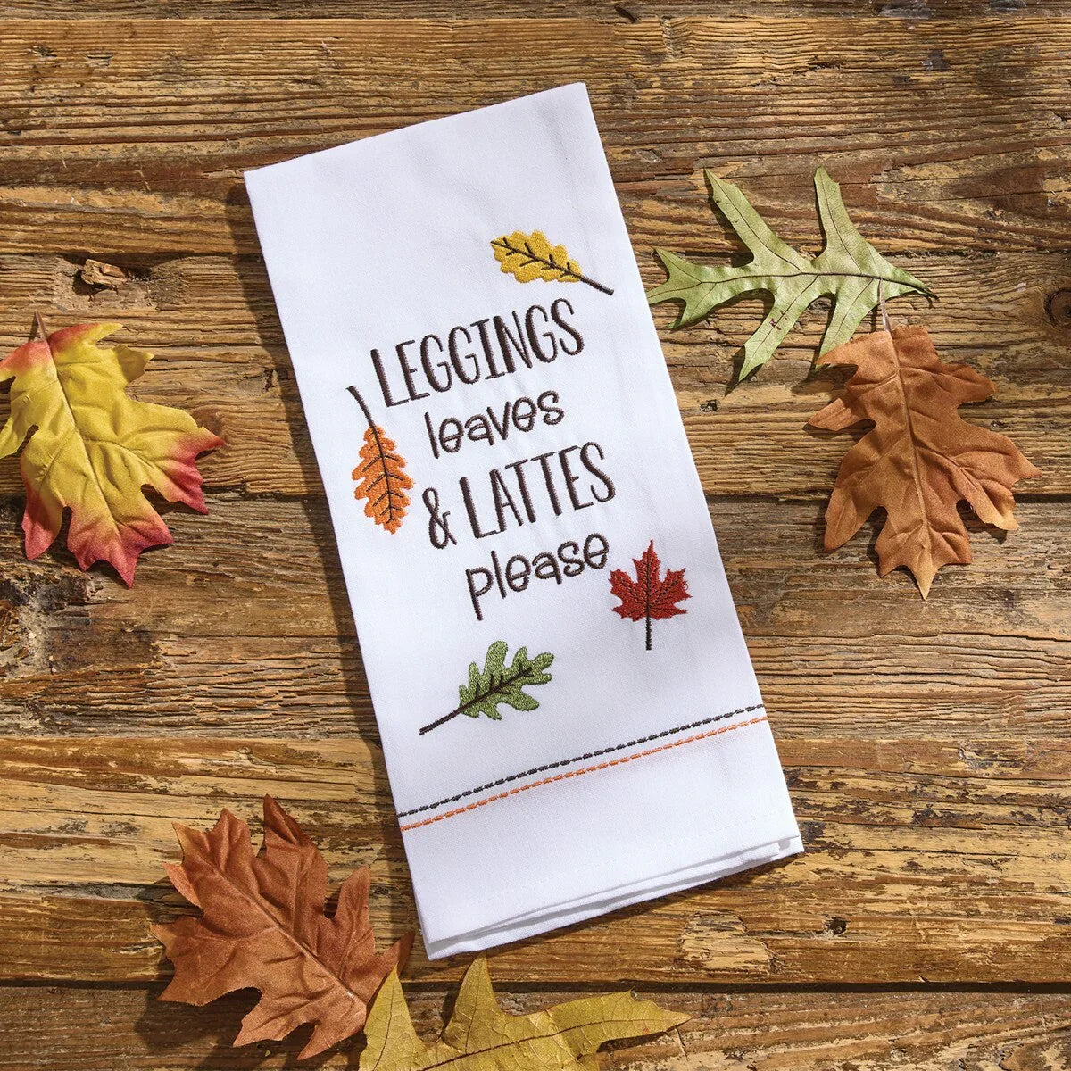 White towel with autumn-themed text and leaf designs on a wooden surface with fall leaves - Your Western Decor