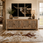 Handcrafted carved wood farmhouse sideboard staged in a bright Western farmhouse interior with oversized longhorn canvas wall art above and cowhide-inspired area rug below, featuring rustic wood textures, shiplap walls, warm neutral tones, and luxury ranch decor styling.