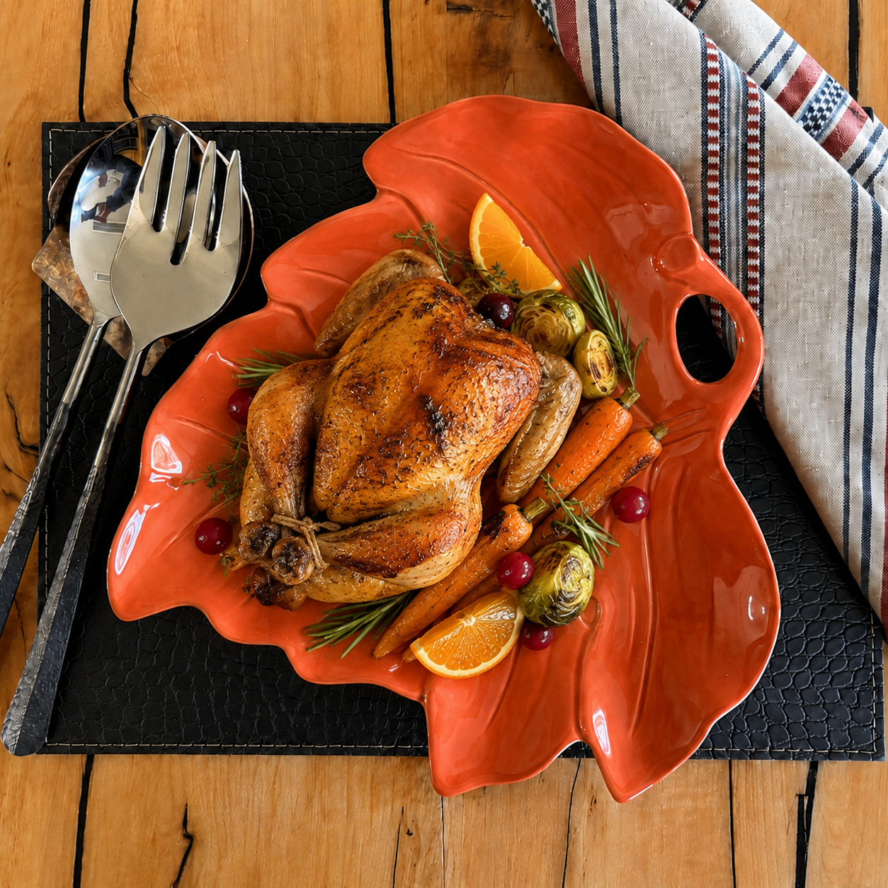 An orange ceramic leaf-shaped serving platter holding a roasted whole chicken with carrots, cranberries, rosemary, and orange slices, set on a black placemat over a wooden table with silverware and a striped napkin.