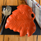 A glossy ceramic leaf-shaped serving platter in a warm orange-red glaze, styled on a black textured placemat with silver cutlery and a striped napkin on a wooden table.