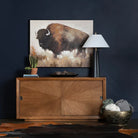 Wooden dresser with a large painting of a bison on a dark wall.