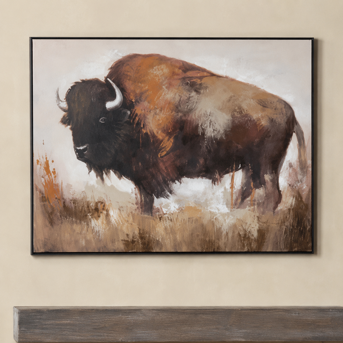 A framed canvas painting of a bison standing in a grassy field, displayed on a beige wall above a dark wooden surface. The painting features earthy brown, black, and beige tones with a black frame.