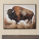 A framed canvas painting of a bison standing in a grassy field, displayed on a beige wall above a dark wooden surface. The painting features earthy brown, black, and beige tones with a black frame.