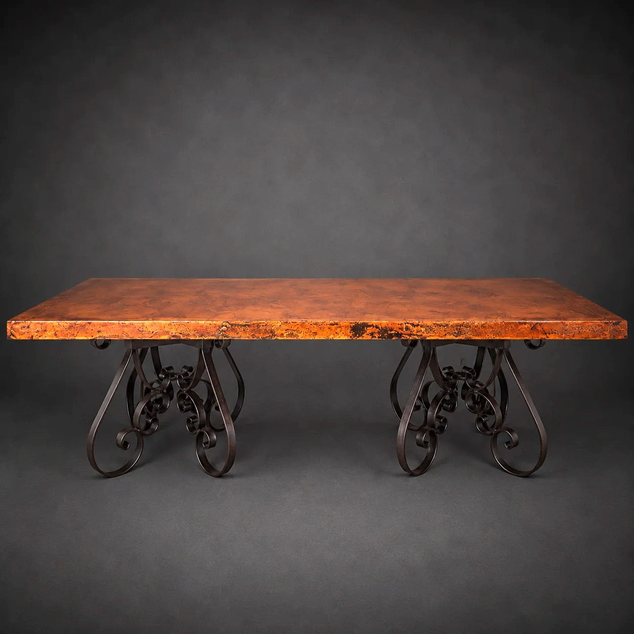 A rustic rectangular table with a distressed copper top and ornate dark metal scrollwork legs, photographed against a dark gray studio background.