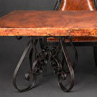 A close-up of an ornate dining table with a rich copper-brown wood top and dark wrought iron scrollwork legs, shown alongside a matching brown leather chair against a dark gray background.