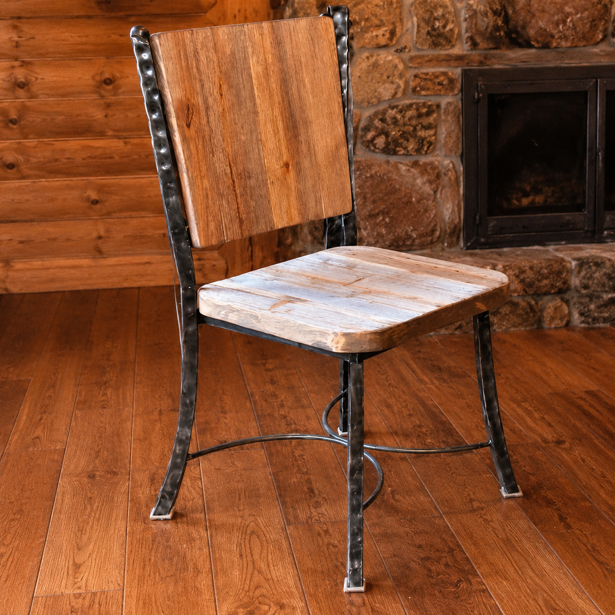 A rustic dining chair with a wood seat and backrest, supported by a dark iron frame with hammered details, set in a cabin-style room with a wood floor and stone fireplace.