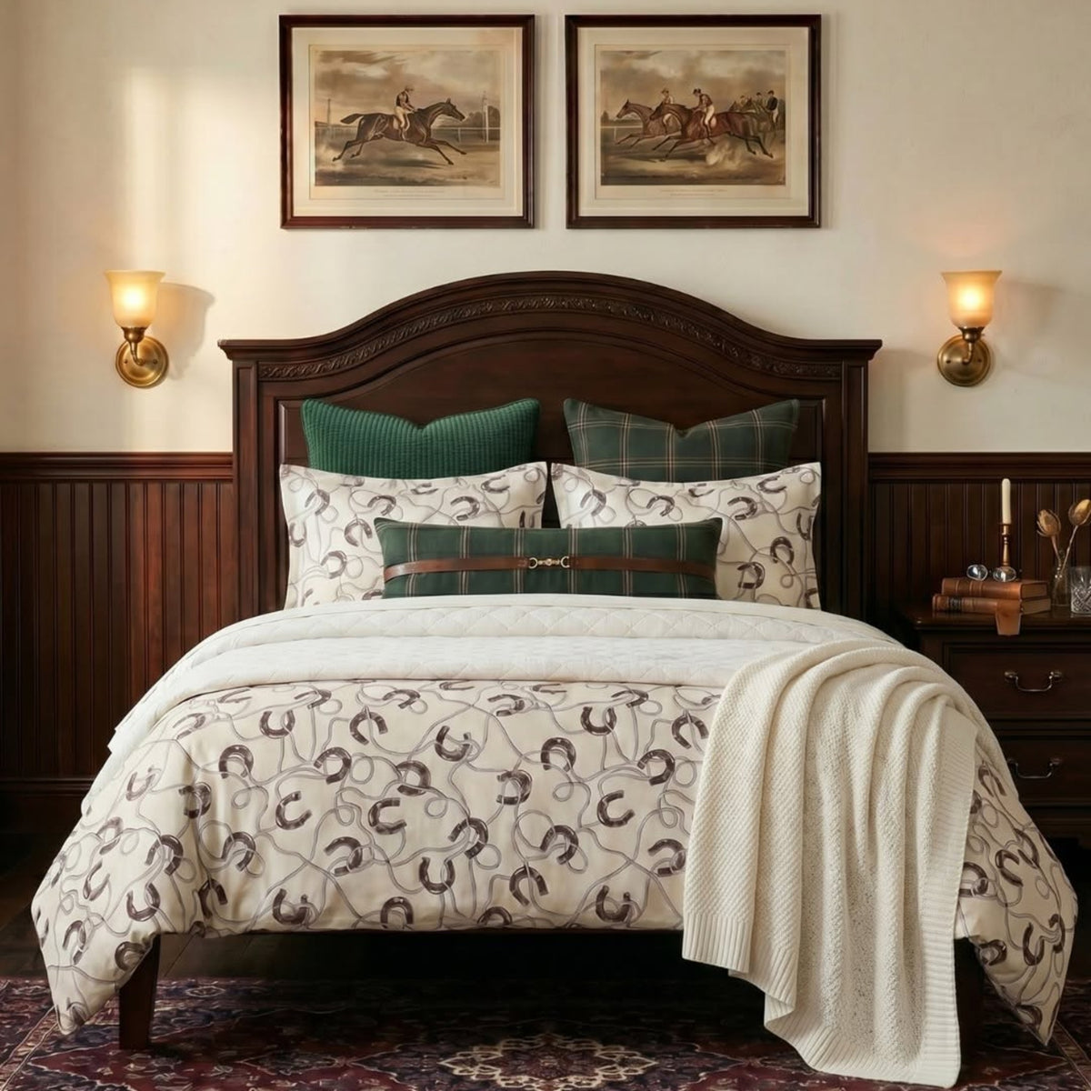 A styled bedroom scene featuring a bed dressed in a cream comforter with a brown horseshoe pattern, matching pillows, green accent pillows, a dark wood headboard and nightstand, wall sconces, framed horse paintings, and a patterned rug.