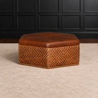 A brown leather ottoman with a diamond-patterned lower skirt and brass nailhead trim, set on a beige carpet in front of a black paneled wall.