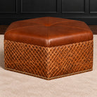 A hexagonal brown leather ottoman with a tufted top and a quilted diamond-patterned base, finished with nailhead trim, shown on a beige carpet in front of a dark paneled wall.