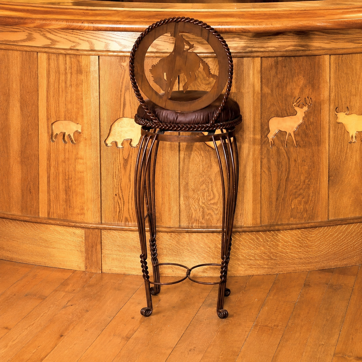 A rustic bar stool with a dark brown leather cushioned seat and a circular backrest featuring a cowboy silhouette, set in front of a curved wooden bar with animal inlays.