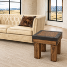 A rustic wooden end table with a dark metal top frame sits next to a beige leather sofa in a bright living room with large windows showing a mountain landscape.