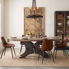 An industrial-style round dining table with a dark metal pedestal base and a light wood top, surrounded by six brown leather chairs with black metal legs, set in a styled dining room.