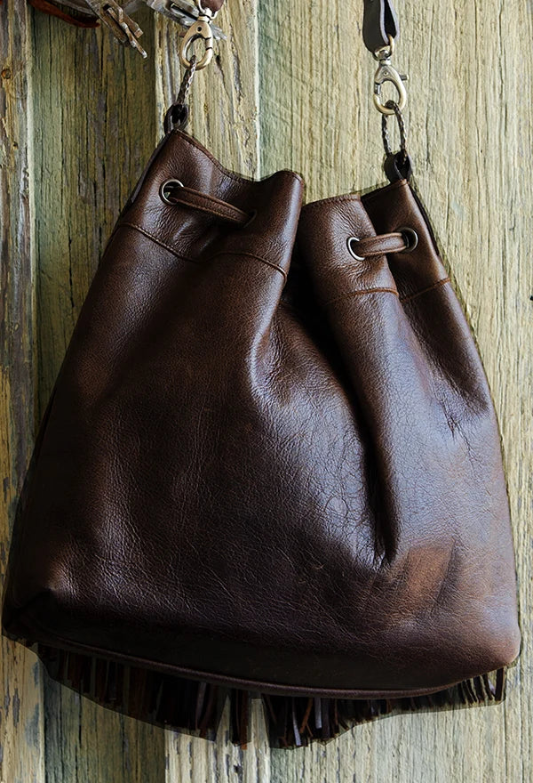 A dark brown leather bucket bag with fringe detailing along the bottom edge, hanging against a rustic wooden plank background.