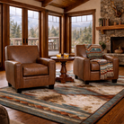 Saddle brown full-grain leather club chairs styled in a luxury mountain lodge living room, set on a Southwestern area rug with large wood-framed windows, exposed beam ceilings, stone fireplace, and whiskey glasses on a round wood side table.