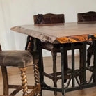A rustic wooden bar height dining table with a live edge, paired with dark brown chairs featuring twisted wooden legs and dark upholstery, set in a dining room.