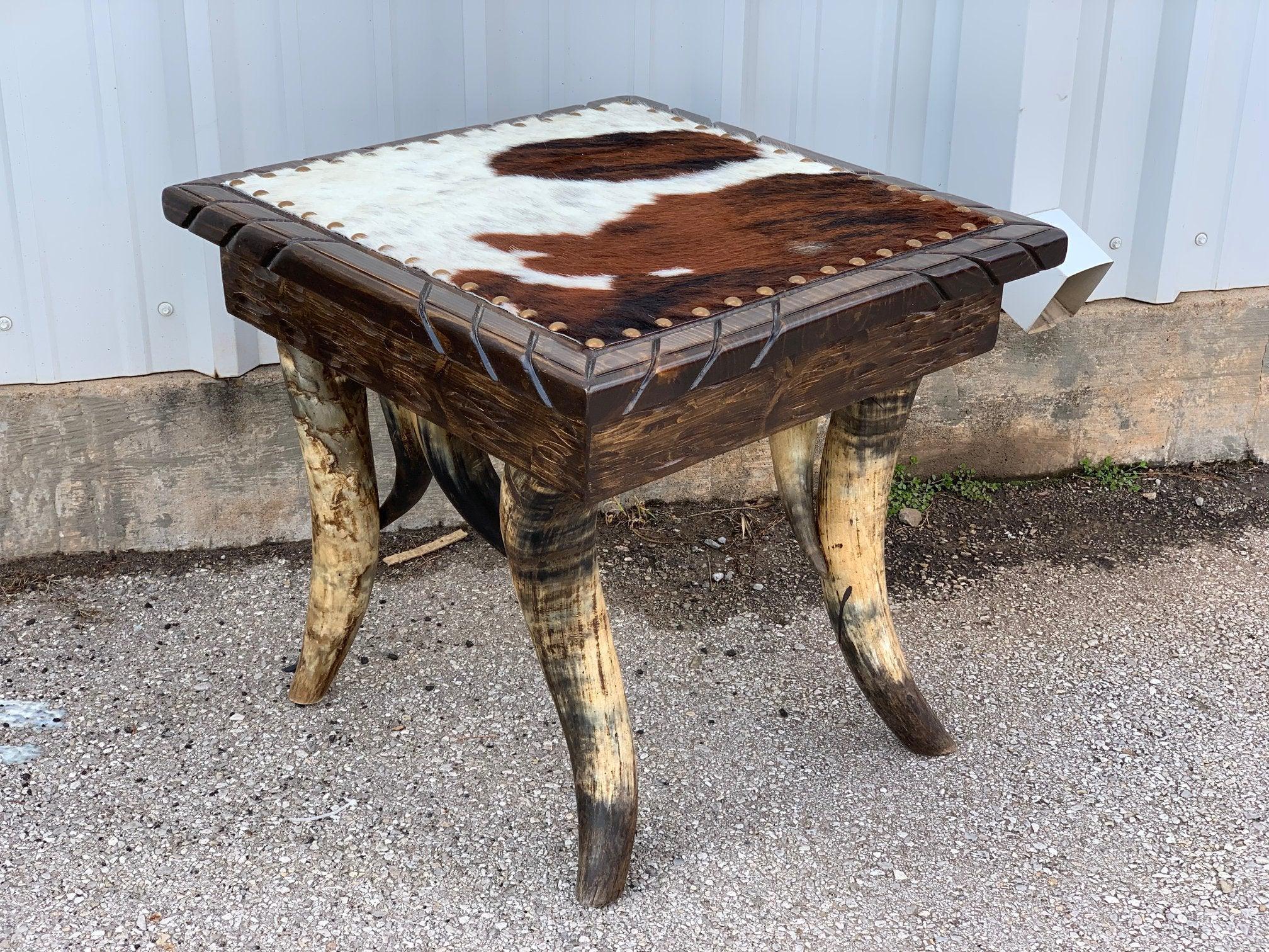 Western end table with carved wood top covered with tri-color cowhide upholstery and nail trim, standing on steer horn legs