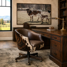 A rustic home office scene featuring a brown and black cowhide accent chair next to a dark wood desk, with a large framed print of a Longhorn cow and calf on the wall behind.