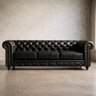 A classic black leather Chesterfield sofa with tufted button detailing, rolled arms, brass nailhead trim, and dark wooden legs, positioned against a textured concrete wall and floor.