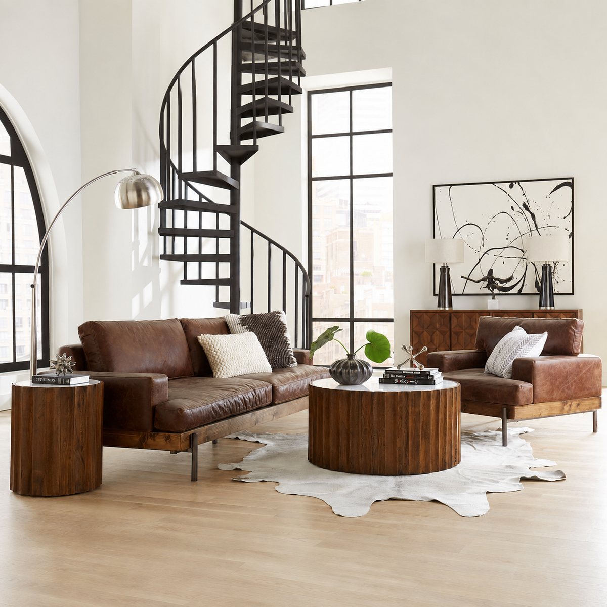 A styled living room featuring a brown leather sofa and matching armchair with wooden bases, arranged around a round wooden coffee table on a white cowhide rug. A black spiral staircase, large arched windows, and abstract wall art are visible in the background.