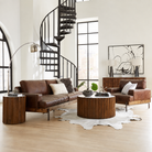 A styled living room featuring a brown leather sofa and matching armchair with wooden bases, arranged around a round wooden coffee table on a white cowhide rug. A black spiral staircase, large arched windows, and abstract wall art are visible in the background.