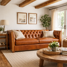 A warm brown leather Chesterfield sofa in a styled living room with white walls, exposed wooden ceiling beams, two framed landscape prints, a side table with a lamp, a round wooden coffee table, and a woven rug.