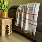 A grey-colored wool throw blanket with a multicolor plaid pattern (pink, blue, orange, and blue stripes) is draped over the back of a dark grey sofa. The scene is styled with a carved wooden side table holding a potted snake plant, set on a light wood floor against a muted olive-green wall.