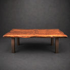 A rustic live-edge wooden table with a rich, warm brown grain pattern and four sturdy dark wrought iron legs, photographed against a dark gray studio background.