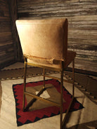 Brown leather bar stool with gold frame on a patterned rug against a wooden wall - Your Western Decor
