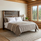 A rustic wooden cedar bed frame with a slatted headboard, dressed in white and taupe bedding, set in a cozy log-cabin style bedroom with a window showing pine trees outside.