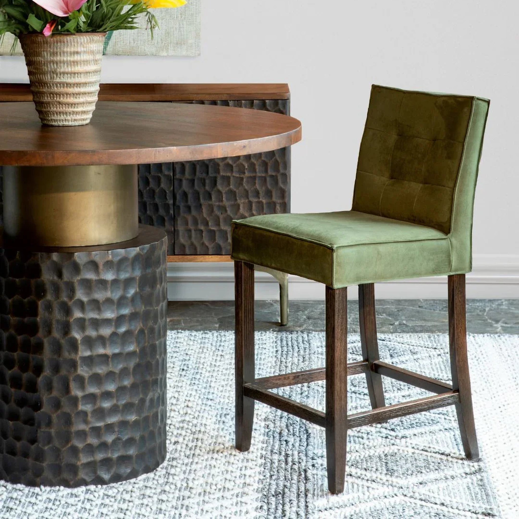 An olive green suede counter stool with a tufted backrest and dark wood legs, positioned next to a round wooden dining table with a hammered metal base in a styled interior setting.