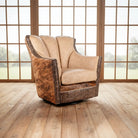 A swivel armchair with a beige suede backrest and brindle cowhide sides, featuring dark metal nailhead trim, set on a light wood floor in front of shoji-style window panels.
