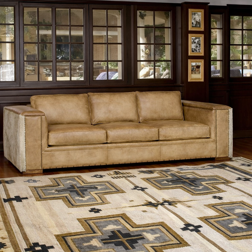 Palomino tan leather sofa with nailhead trim positioned partially over a Southwestern geometric area rug in a warm ranch-style living room with wood floors and large windowed wall.