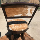 A rustic swivel bar stool with a dark brown iron frame, featuring a curved backrest and seat upholstered in beige and white cowhide with a wide black 