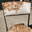 A close-up of a chair upholstered in cowhide, showing a brown and white mottled pattern on the backrest and seat cushion, with dark metal legs and frame.